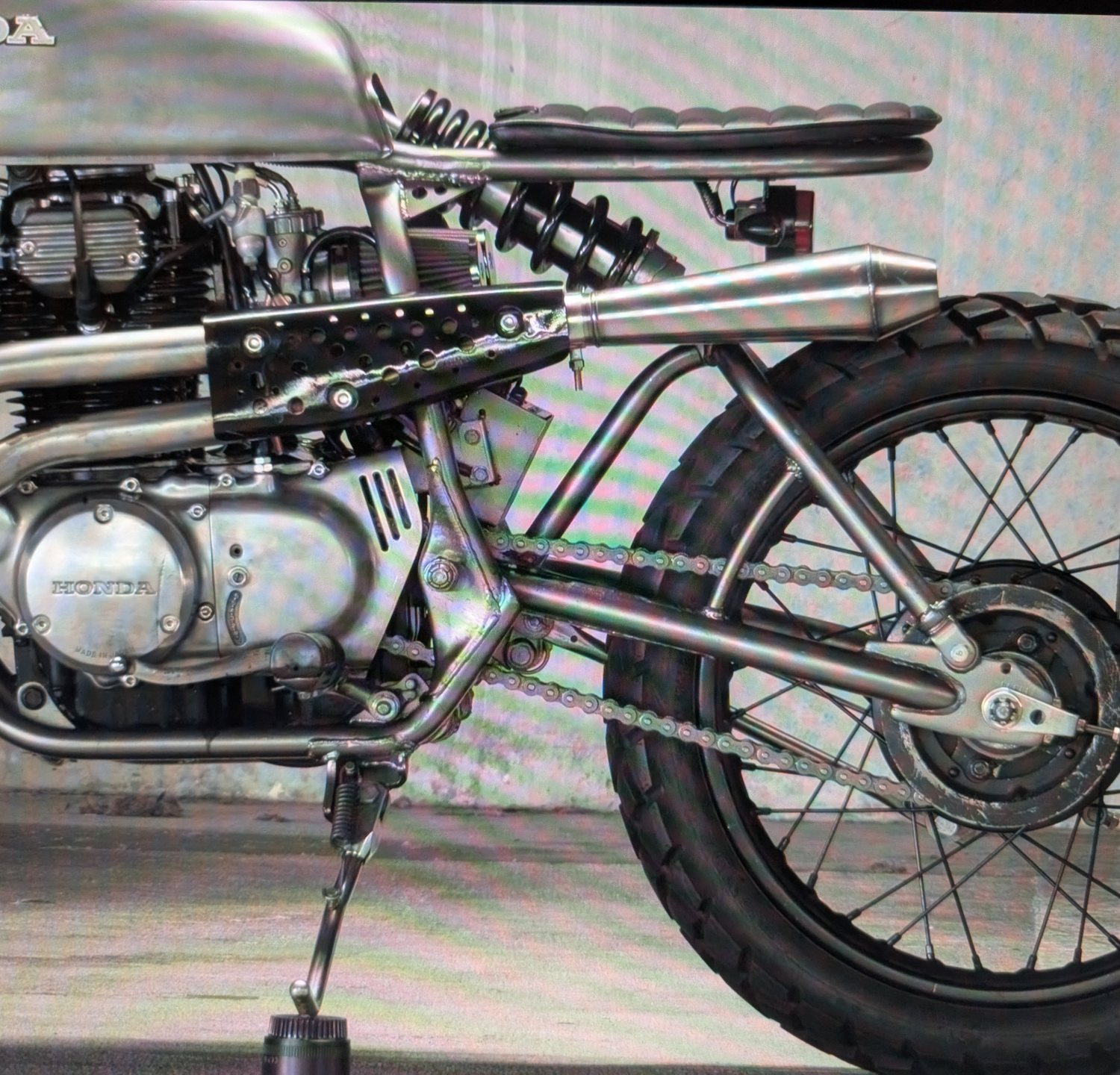 Raw metal Honda cafe racer, close on the engine, perforated exhaust shield and knobby rear tyre