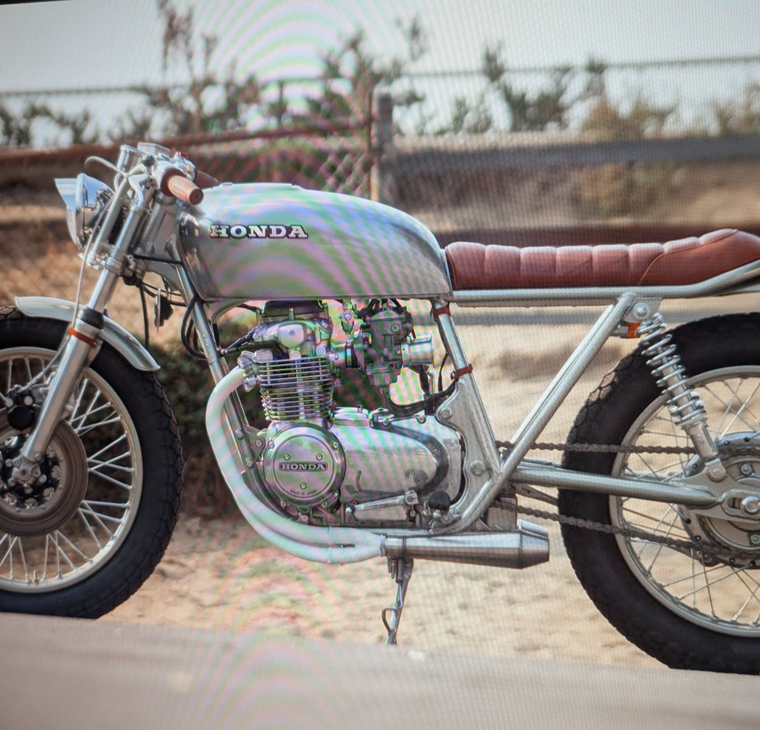 Silver Honda CB550 cafe racer with a tan seat, parked in desert scrub